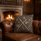 A square decorative throw pillow with a floral tapestry pattern and crossed swords, resting on a brown leather tufted armchair in front of a warm fireplace.