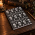 A spiral-bound notebook with a black cover decorated with a repeating white skull and damask floral pattern, displayed on a rustic wooden table next to a lit candle, an antique book, and a gold pen.