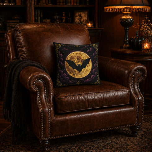 A square decorative throw pillow with a gothic bat-and-moon design, resting on a brown leather armchair in a cozy, dimly lit room with a lamp and bookshelves in the background.