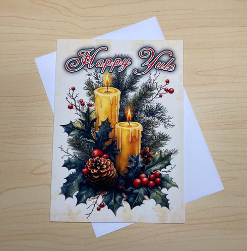A square Christmas greeting card with a white envelope, placed on a light wood surface. The card features an illustration of two lit yellow candles, holly leaves, red berries, and pinecones, with the text 'Happy Yule' in stylized red and blue lettering.