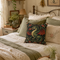 A decorative throw pillow featuring a detailed green dragon illustration with red wings and floral details, styled on a bed with other sage green and cream pillows in a cozy, rustic bedroom setting.