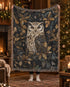A person holds up a large woven throw blanket featuring a detailed owl and holly pattern in dark blue, gold, and cream tones, with fringed edges, set against a cosy Christmas living room background.