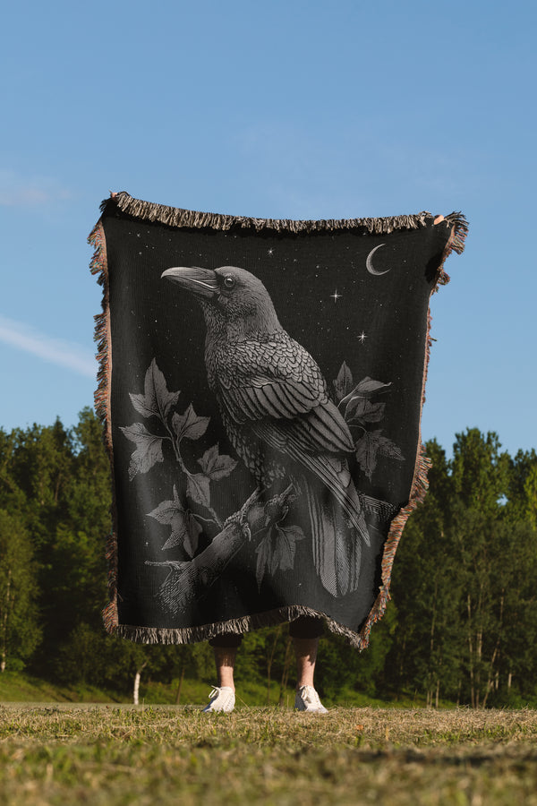 A black woven throw blanket with a detailed grayscale raven and moon design, held up outdoors by a person whose legs and feet are visible below the blanket against a background of trees and blue sky.