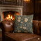 A square decorative throw pillow with a floral pattern in muted blue, beige, and green, resting on a brown leather Chesterfield sofa in front of a warm fireplace.
