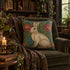 A decorative square throw pillow featuring a cream-colored rabbit on a teal and pink floral background, styled on an olive green armchair in a cozy, book-filled room.