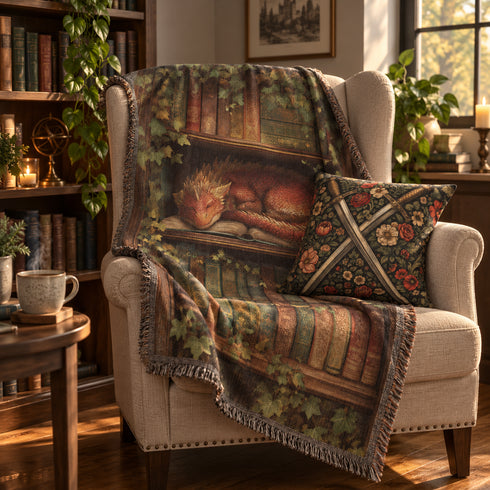 A cosy living room scene with a beige armchair draped in a printed throw blanket featuring a sleeping red dragon on bookshelves, alongside a matching floral accent pillow with crossed medieval swords. The room includes bookshelves, plants, and warm window light.