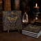 A stack of soapstone absorbent coasters with an ornate gold and purple moth design, displayed on a dark wooden table with a lit candle, crystal, and antique books in the background.