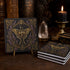 A stack of soapstone absorbent coasters with an ornate gold and purple moth design, displayed on a dark wooden table with a lit candle, crystal, and antique books in the background.