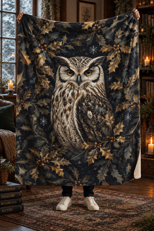 A person holds up a large fleece blanket featuring a detailed illustration of an owl surrounded by holly leaves and snowflakes on a dark background, set in a cozy, warmly lit room.