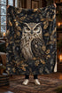 A person holds up a large fleece blanket featuring a detailed illustration of an owl surrounded by holly leaves and snowflakes on a dark background, set in a cozy, warmly lit room.