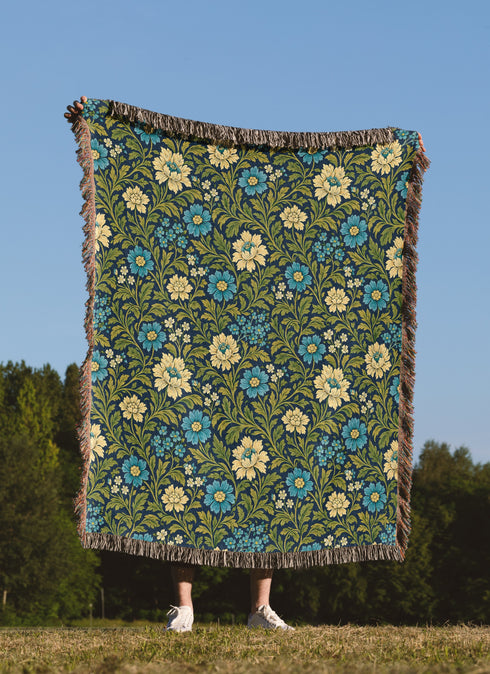 A person standing outdoors in a grassy field under a clear blue sky holds up a large woven blanket or tapestry with a floral pattern. The blanket has a dark green background with blue and cream flowers, green leaves, and fringed edges.