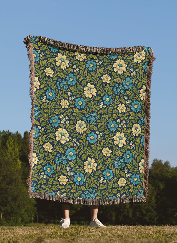 A person standing outdoors in a grassy field under a clear blue sky holds up a large woven blanket or tapestry with a floral pattern. The blanket has a dark green background with blue and cream flowers, green leaves, and fringed edges.