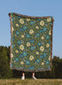A person standing outdoors in a grassy field under a clear blue sky holds up a large woven blanket or tapestry with a floral pattern. The blanket has a dark green background with blue and cream flowers, green leaves, and fringed edges.